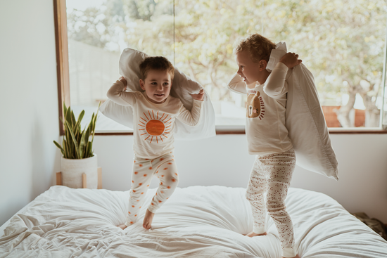Two kids in comfortable Rising Sun and Dreamy Lion Tiny Otters children's pajamas enjoying a pillow fight on a bed
